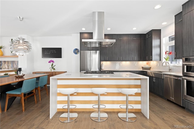 kitchen with appliances with stainless steel finishes, a kitchen island, island exhaust hood, a kitchen breakfast bar, and light hardwood / wood-style flooring