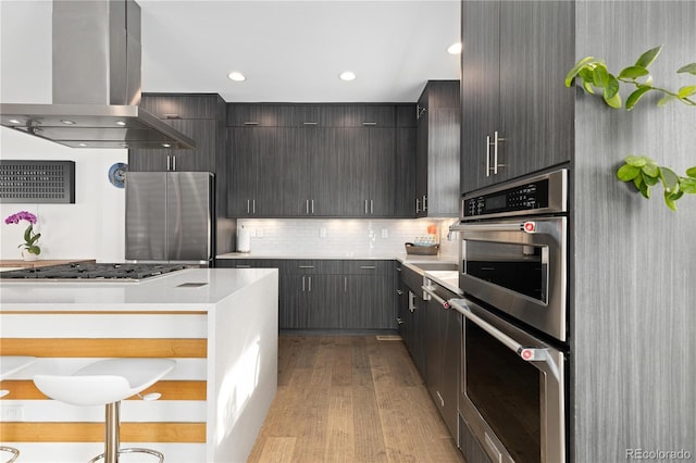 kitchen featuring a breakfast bar, island range hood, light wood-type flooring, backsplash, and stainless steel appliances