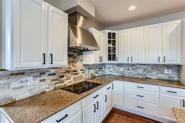 kitchen with wall chimney range hood, glass insert cabinets, black electric stovetop, and white cabinetry
