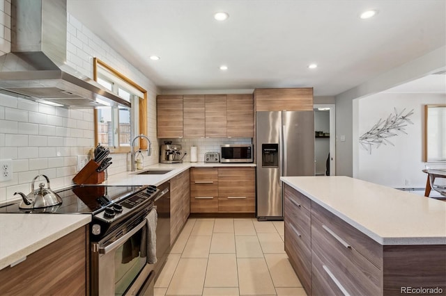 kitchen featuring appliances with stainless steel finishes, wall chimney exhaust hood, a sink, tasteful backsplash, and brown cabinets