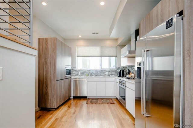 kitchen with appliances with stainless steel finishes, light hardwood / wood-style floors, white cabinetry, and wall chimney exhaust hood