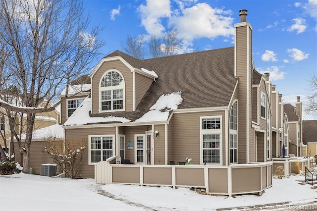 snow covered rear of property with a shingled roof, a chimney, and central air condition unit