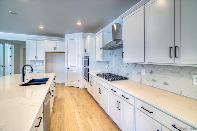 kitchen with wall chimney range hood, light wood-style flooring, stainless steel appliances, white cabinetry, and a sink