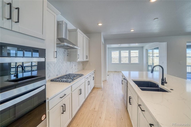 kitchen featuring a sink, light stone counters, appliances with stainless steel finishes, white cabinets, and wall chimney range hood