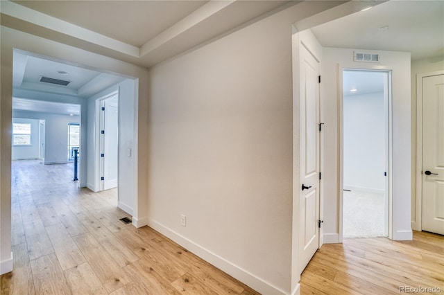 hall with a tray ceiling, visible vents, baseboards, and light wood finished floors