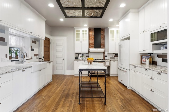 kitchen with white appliances, light stone counters, wall chimney exhaust hood, white cabinetry, and sink