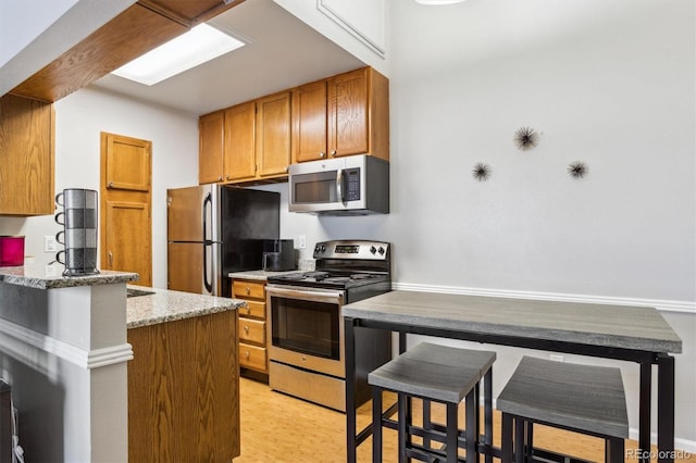 kitchen with brown cabinetry, appliances with stainless steel finishes, a breakfast bar, light stone countertops, and light wood-type flooring