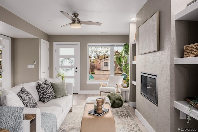 living area with ceiling fan, a glass covered fireplace, light wood-style floors, and baseboards