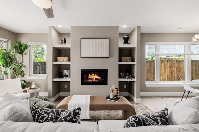 living room featuring a glass covered fireplace, wood finished floors, baseboards, ceiling fan, and built in features