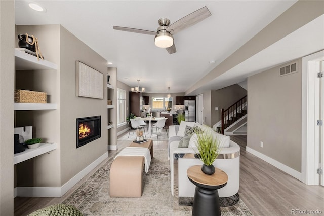 living room with built in features, a ceiling fan, baseboards, stairs, and light wood-style floors