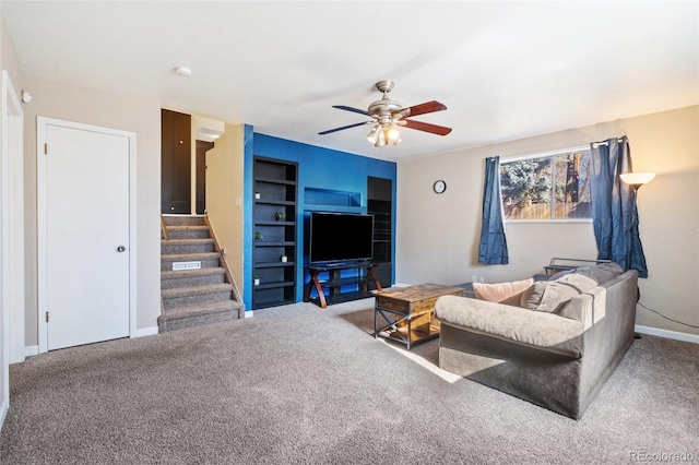 carpeted living area with built in shelves, ceiling fan, stairway, and baseboards