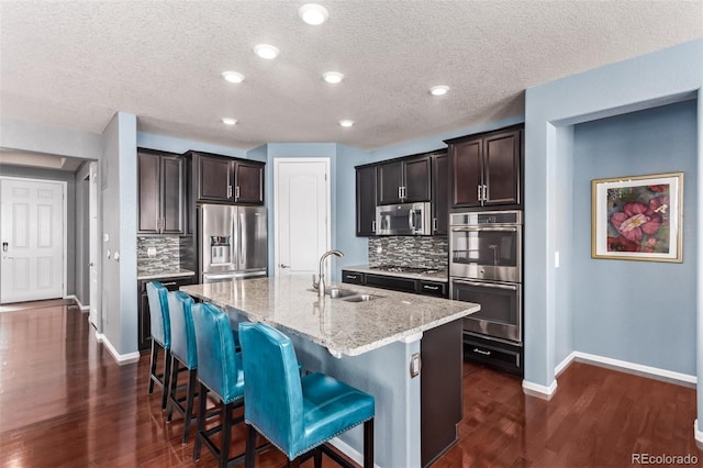 kitchen featuring a center island with sink, appliances with stainless steel finishes, dark hardwood / wood-style flooring, light stone countertops, and sink