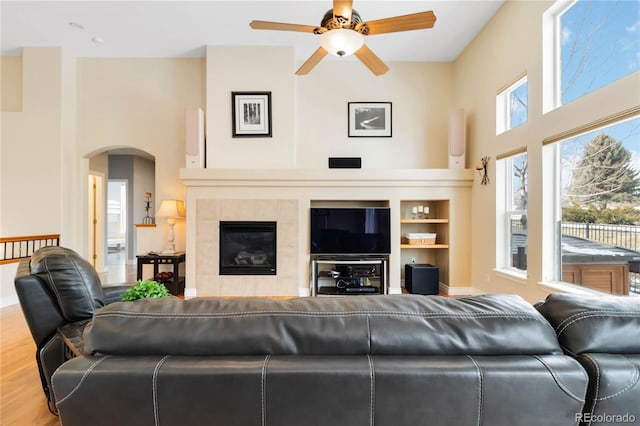 living room featuring arched walkways, a high ceiling, a tiled fireplace, a ceiling fan, and wood finished floors