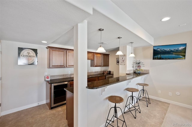 kitchen with a breakfast bar, hanging light fixtures, dark stone countertops, kitchen peninsula, and beverage cooler