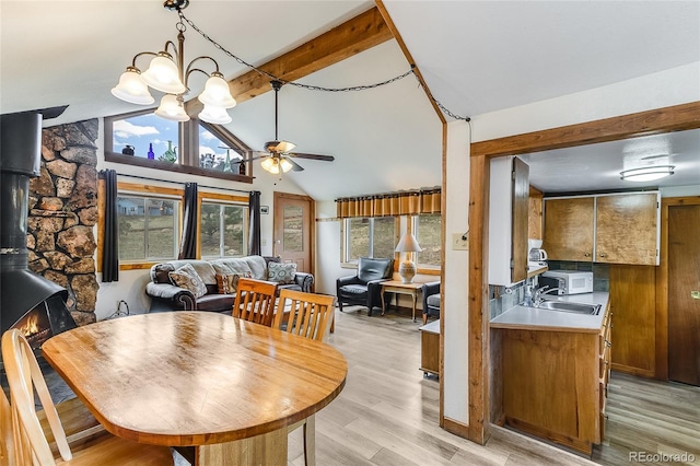 dining space featuring a wood stove, light wood-type flooring, vaulted ceiling with beams, ceiling fan with notable chandelier, and sink