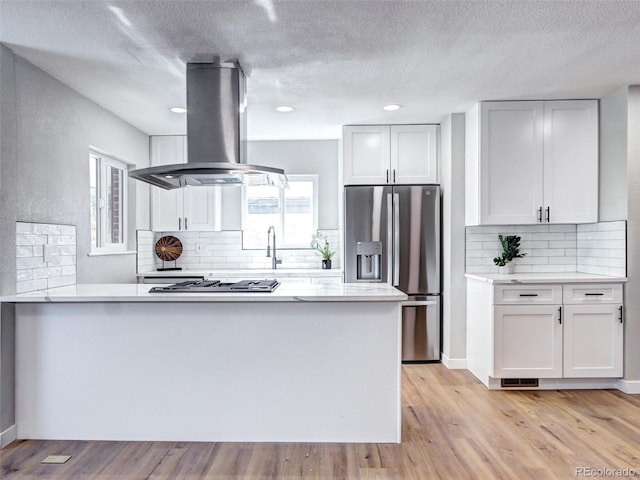 kitchen featuring light wood finished floors, a peninsula, island exhaust hood, stainless steel appliances, and light countertops