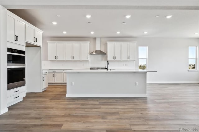 kitchen featuring stainless steel double oven, light wood finished floors, wall chimney exhaust hood, and recessed lighting