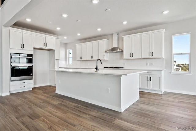 kitchen with wall chimney range hood, plenty of natural light, a center island with sink, and white cabinetry