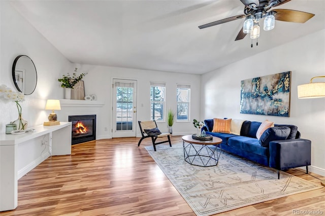 living area featuring a ceiling fan, baseboards, vaulted ceiling, light wood finished floors, and a glass covered fireplace