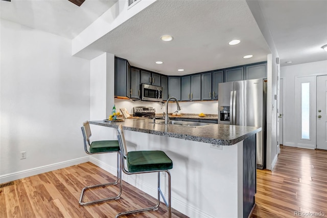 kitchen featuring stainless steel appliances, a peninsula, wood finished floors, a sink, and dark countertops