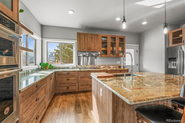 kitchen with stainless steel appliances, wood finished floors, brown cabinetry, light stone countertops, and under cabinet range hood