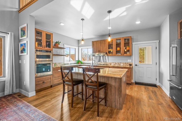 kitchen with stainless steel double oven, high end refrigerator, wood finished floors, brown cabinets, and an island with sink