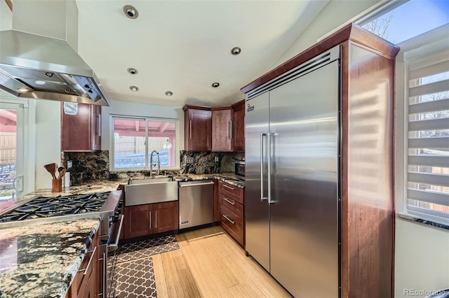 kitchen featuring island exhaust hood, light wood finished floors, stainless steel appliances, a sink, and dark stone countertops