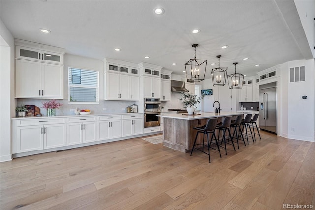 kitchen with wall chimney range hood, an island with sink, pendant lighting, white cabinetry, and appliances with stainless steel finishes