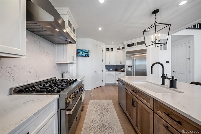 kitchen with wall chimney range hood, sink, light wood-type flooring, white cabinets, and high end appliances