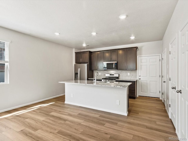 kitchen with dark brown cabinetry, decorative backsplash, light wood-style floors, stainless steel appliances, and a kitchen island with sink