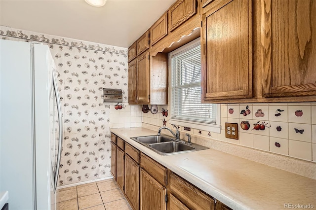 kitchen featuring a sink, freestanding refrigerator, brown cabinetry, wallpapered walls, and light tile patterned floors