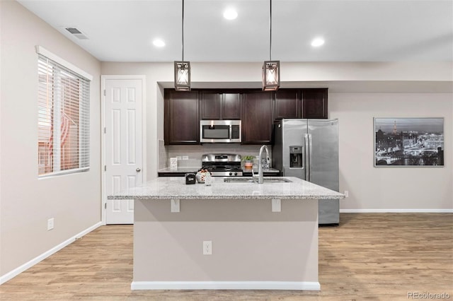 kitchen with sink, an island with sink, appliances with stainless steel finishes, dark brown cabinets, and light stone counters