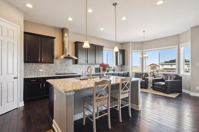 kitchen with wall chimney range hood, backsplash, light stone countertops, and a kitchen island with sink