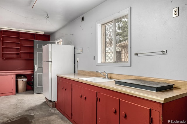 kitchen with light countertops, unfinished concrete flooring, red cabinetry, a sink, and freestanding refrigerator