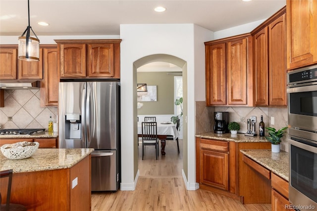 kitchen featuring light stone countertops, under cabinet range hood, brown cabinets, appliances with stainless steel finishes, and arched walkways