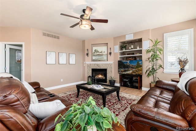 living room featuring baseboards, a warm lit fireplace, light wood-style floors, visible vents, and ceiling fan