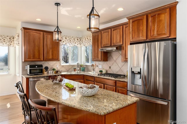 kitchen featuring light stone counters, under cabinet range hood, brown cabinets, stainless steel appliances, and a sink