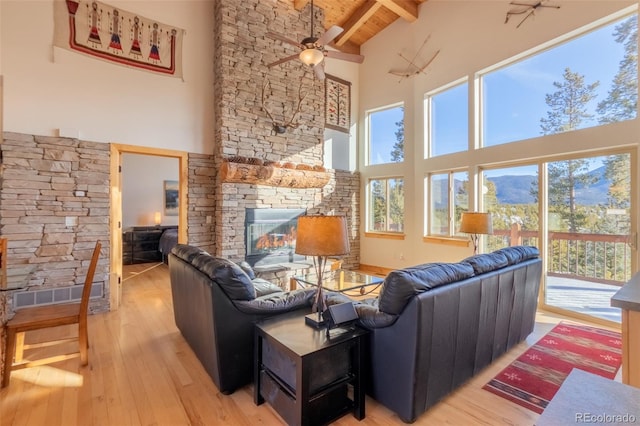 living room featuring beamed ceiling, a stone fireplace, a healthy amount of sunlight, and light wood-type flooring