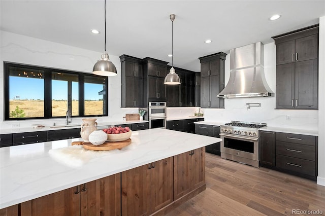 kitchen with appliances with stainless steel finishes, light hardwood / wood-style floors, wall chimney exhaust hood, decorative light fixtures, and a center island