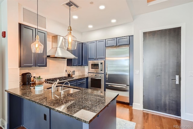 kitchen featuring wall chimney range hood, dark stone countertops, hanging light fixtures, built in appliances, and kitchen peninsula