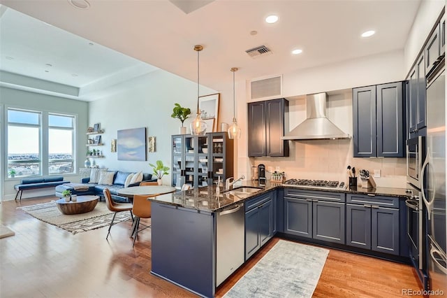 kitchen featuring wall chimney exhaust hood, sink, dark stone countertops, appliances with stainless steel finishes, and pendant lighting