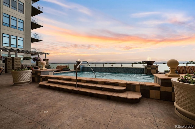 pool at dusk featuring a hot tub, pool water feature, and a water view