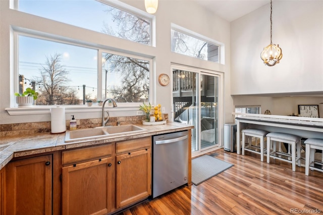 kitchen featuring brown cabinets, wood finished floors, decorative light fixtures, stainless steel dishwasher, and a sink