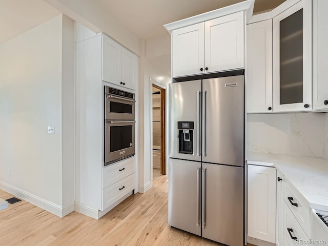 kitchen featuring appliances with stainless steel finishes, light wood-style floors, white cabinetry, glass insert cabinets, and light stone counters