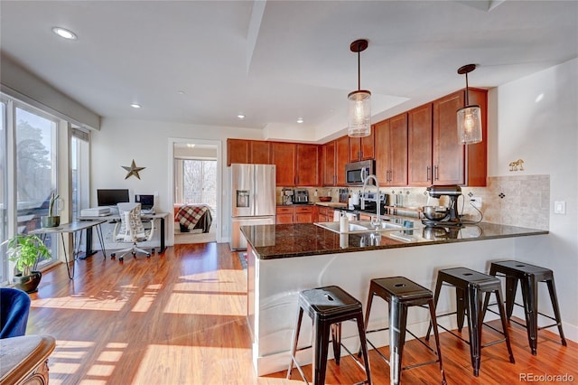 kitchen with appliances with stainless steel finishes, sink, dark stone countertops, hanging light fixtures, and light hardwood / wood-style flooring