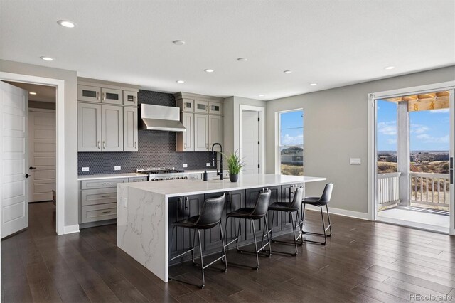 kitchen featuring gray cabinets, a kitchen island with sink, a kitchen bar, tasteful backsplash, and wall chimney exhaust hood