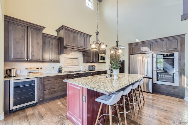 kitchen with wine cooler, hanging light fixtures, a kitchen island with sink, stainless steel appliances, and a high ceiling