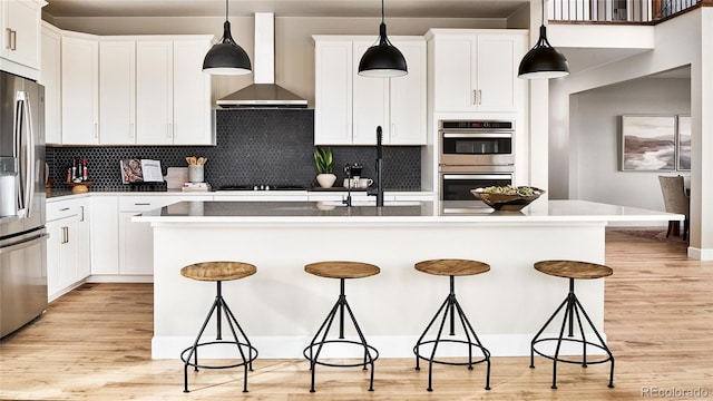 kitchen with wall chimney exhaust hood, decorative light fixtures, a kitchen island with sink, and appliances with stainless steel finishes