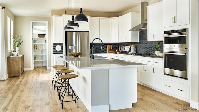 kitchen featuring light wood-type flooring, wall chimney exhaust hood, stainless steel appliances, sink, and pendant lighting