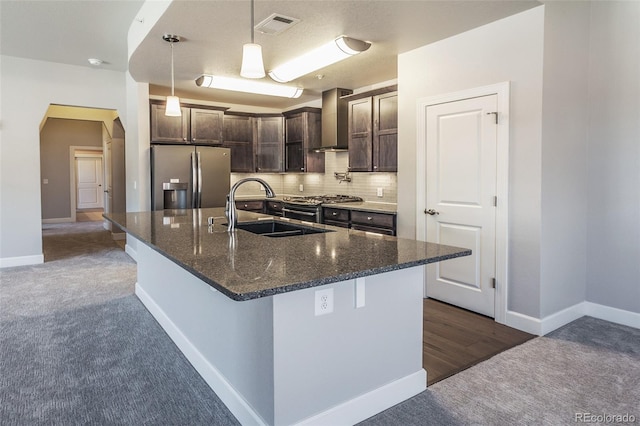 kitchen featuring wall chimney exhaust hood, stainless steel fridge with ice dispenser, an island with sink, dark stone counters, and dark brown cabinets